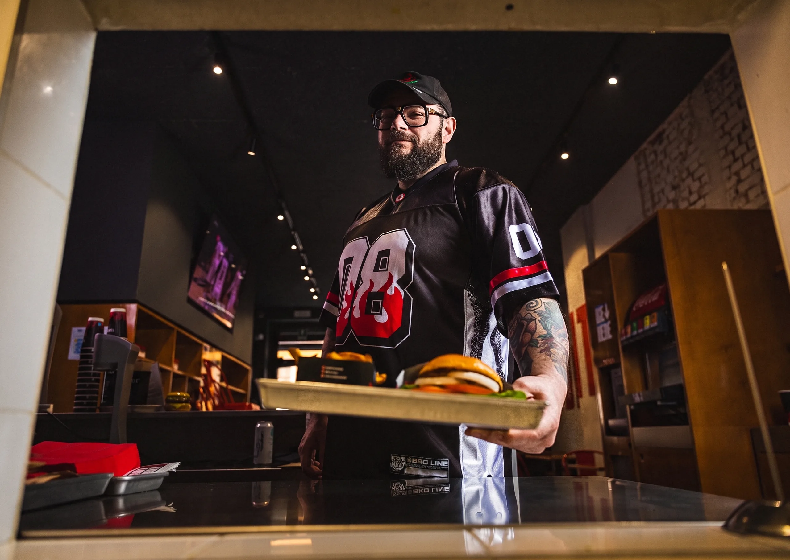 Mocho at the pass of a Meat Crew store in the 08 jersey, serving a tray with a burger and fries