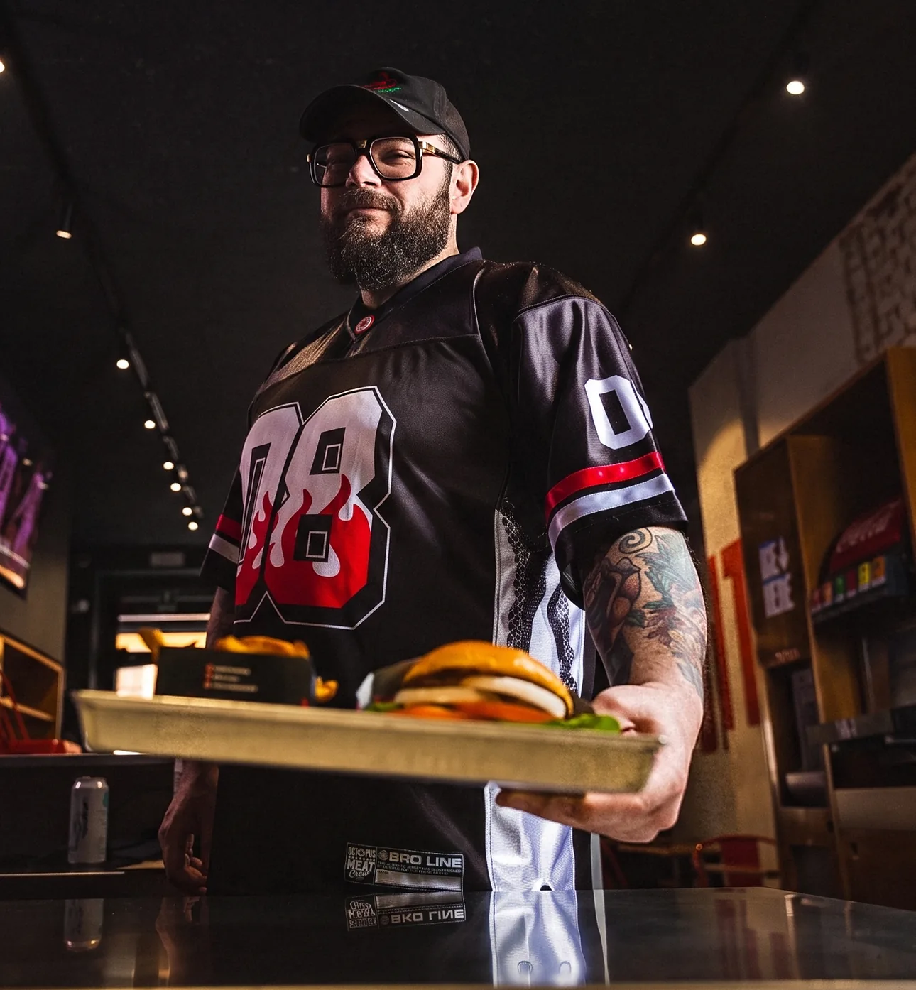 Mocho at the pass of a Meat Crew store in the 08 jersey, serving a tray with a burger and fries
